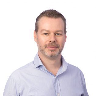 A man with light skin, short brown hair, and a trimmed beard is wearing a light blue collared shirt and posing against a plain white background. He is looking at the camera with a neutral expression.